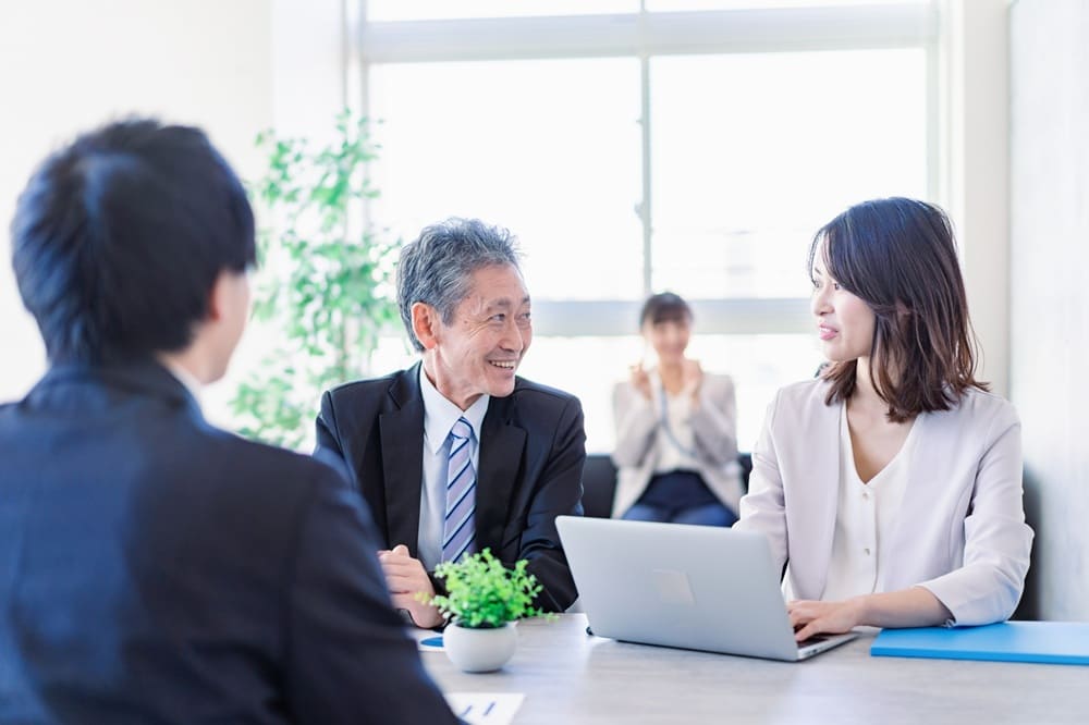 オープンオフィス内でにこやかに話すスーツの男女と、それを手前と奥で見守るスーツの男女