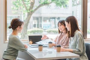 窓際の明るい店内で、それぞれコーヒーを手に女性3人で仕事の打ち合わせ中