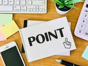 POINTと黒字で書かれたノートを囲むようにスマホや電卓、ノートPCが置かれたデスク