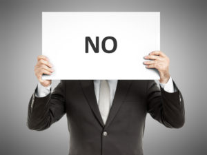 「NO」と書かれたパネルを顔前に掲げているスーツの男性
