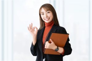 ノートPCを左手にかかえて右手でOKサインを出すビジネスウーマン