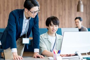 オフィスで先輩男性社員の指導をノートPCを操作しながら受ける後輩の男性社員